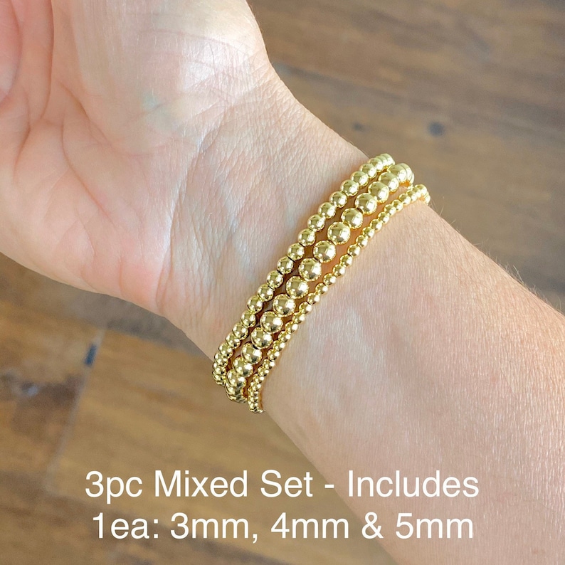 May include: A set of three gold bead bracelets, each with a different size bead: 3mm, 4mm, and 5mm. The bracelets are stacked on a person's wrist.