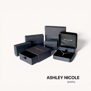 May include: An assortment of black jewelry boxes, some open to reveal a necklace. The boxes are various sizes and shapes, with the brand name "ASHLEY NICOLE" printed on them. The word "jewelry" is below the brand name.