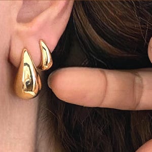Puede incluir: Un par de pendientes de aro de oro en forma de lágrima. Los pendientes están superpuestos, con un aro más pequeño dentro de un aro más grande.