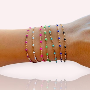 Könnte beinhalten: Ein Set aus acht goldenen Kettenarmbändern mit bunten Perlen. Die Armbänder sind auf dem Handgelenk einer Person gestapelt. Die Perlen sind in verschiedenen Farben, darunter rosa, blau, grün und schwarz.