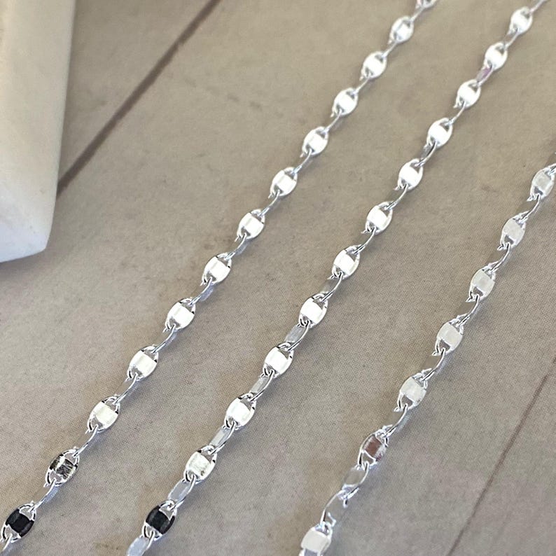 May include: Three silver chains with a delicate oval link design. The chains are laid out in a row on a light grey surface.