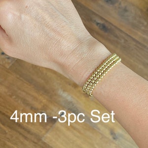 May include: A set of three gold beaded bracelets, each with 4mm beads. The bracelets are stacked on a person's wrist. The text "4mm - 3pc Set" is visible below the bracelets.