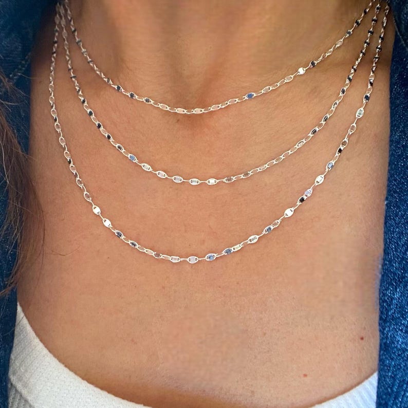 May include: Three silver chain necklaces with a delicate, oval-shaped link design. The necklaces are layered on a person's neck.