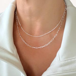 May include: Two silver necklaces layered together. The bottom necklace has a chain link design, while the top necklace is a delicate beaded chain. The necklaces are worn against a white background.