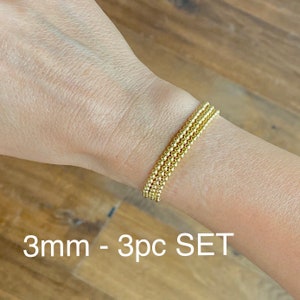 May include: A set of three gold-toned beaded bracelets, each with 3mm beads. The bracelets are stacked on a person's wrist. The text "3mm - 3pc SET" is visible below the bracelets.