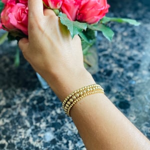 May include: A gold beaded bracelet with three strands of different sized beads. The bracelet is worn on a person's wrist.