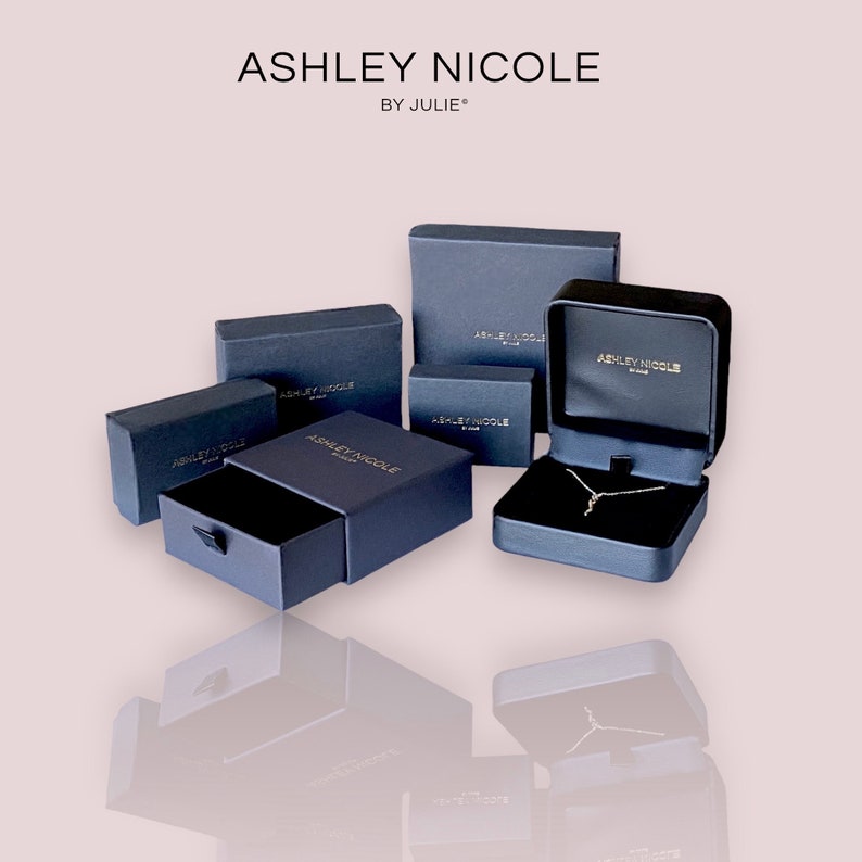 May include: Several black gift boxes with gold lettering that reads "Ashley Nicole by Julie". The boxes are arranged in a pyramid shape on a white surface.
