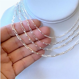 May include: Four delicate silver chain necklaces with rectangular links are displayed. The chains are draped over a white surface, showcasing their reflective quality. The necklaces are of varying lengths, offering layering options.