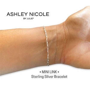 May include: A silver chain bracelet with a clasp closure. The bracelet is on a person's wrist. The text "MINI LINK" and "Sterling Silver Bracelet" is below the bracelet.