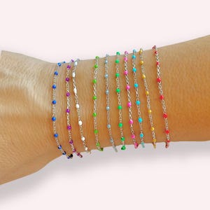 May include: A set of 12 colourful bracelets with small beads on a silver chain. The bracelets are stacked on a person's wrist.