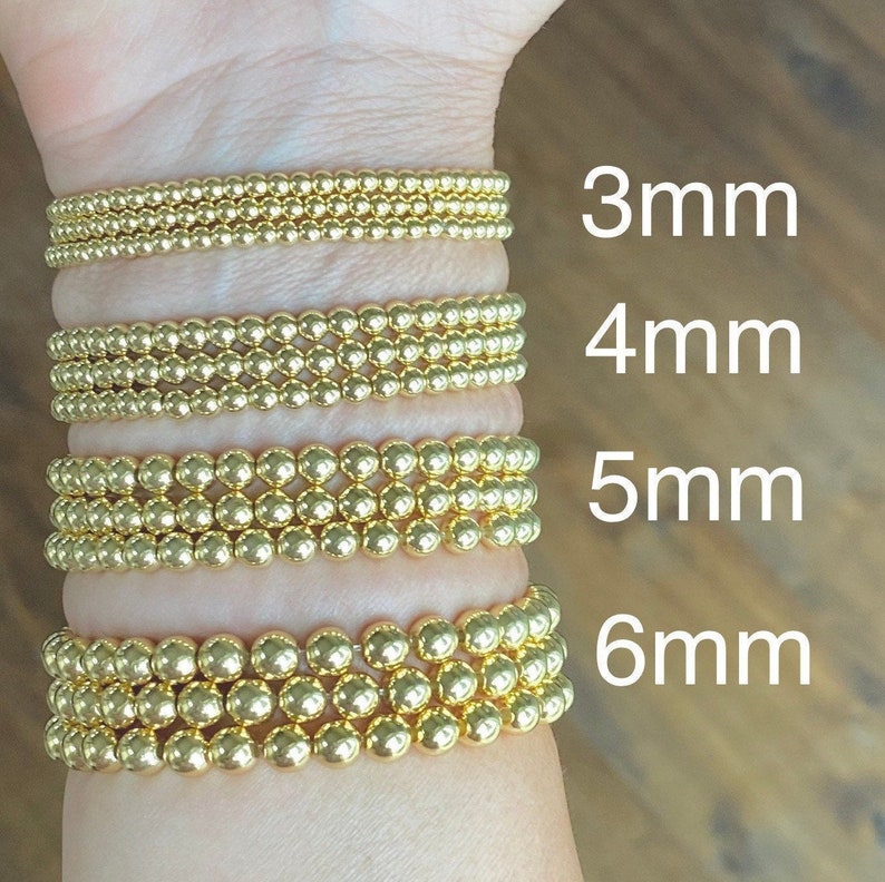 May include: Four gold bracelets with different sized beads. The bracelets are stacked on a person's wrist. The bead sizes are 3mm, 4mm, 5mm, and 6mm.
