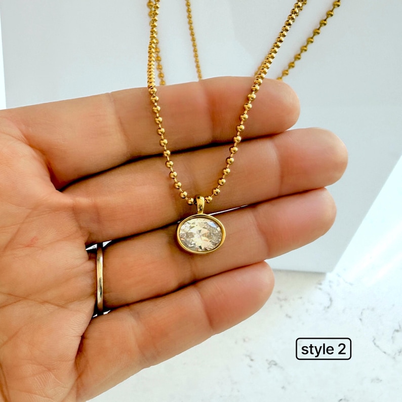 Close-up of oval cubic zirconia pendant on 18K PVD gold 2mm ball chain held in palm showing warm yellow gold tone and sparkling CZ stone detail, style 2