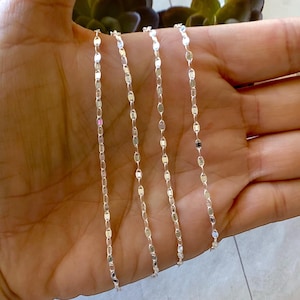 Sparkling silver chains with a delicate, flat oval link design. The chains are laid out in a row on a hand, showing the length and detail of the links.