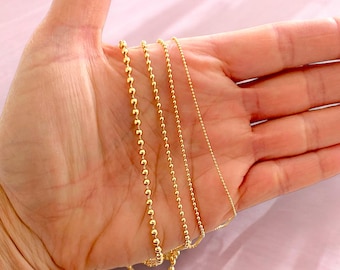 May include: Four gold chains of different lengths and thicknesses, with round beads, are displayed on a hand.