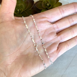 May include: A silver chain necklace with a delicate, flat, oval-shaped link design. The chain is shown lying on a hand.