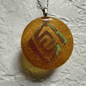 May include: A gold round pendant necklace with a green and pink geometric design. The pendant is made of resin and has a glittery finish.