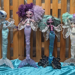 May include: Four mermaid dolls with long, flowing hair and colourful tails. The dolls are wearing different outfits and have different hairstyles. The dolls are all standing on a blue surface.