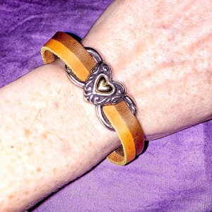 May include: A leather bracelet with a silver heart-shaped clasp. The bracelet features a two-tone design with shades of brown and tan. The clasp has an ornate design with a gold heart in the center. The bracelet is worn on a wrist.