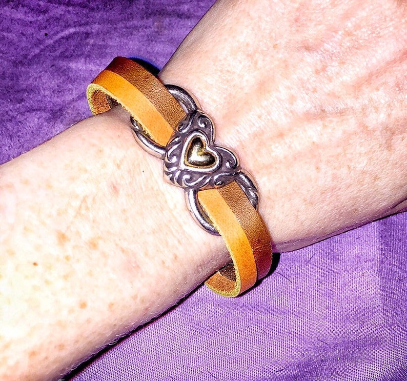 May include: A leather bracelet with a silver heart-shaped clasp. The bracelet features a two-tone design with shades of brown and tan. The clasp has an ornate design with a gold heart in the center. The bracelet is worn on a wrist.