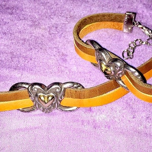 May include: Two leather bracelets with silver-tone heart-shaped clasps. The bracelets feature a layered design with brown, tan, and yellow-orange leather straps. The background is a textured purple fabric.