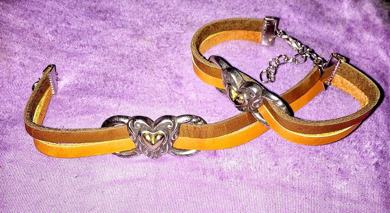 May include: Two leather bracelets with silver-tone heart-shaped clasps. The bracelets feature a layered design with brown, tan, and yellow-orange leather straps. The background is a textured purple fabric.