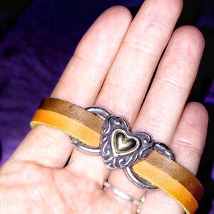 May include: A leather bracelet with a silver heart-shaped centerpiece. The bracelet features a two-tone brown and yellow leather band. The heart has a gold center and ornate silver detailing. The bracelet is shown on a hand.