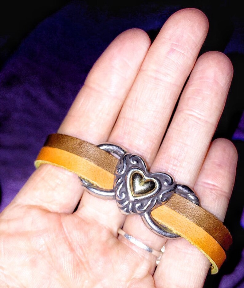 May include: A leather bracelet with a silver heart-shaped centerpiece. The bracelet features a two-tone brown and yellow leather band. The heart has a gold center and ornate silver detailing. The bracelet is shown on a hand.