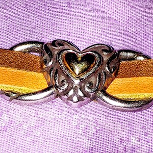 May include: A close-up of a leather bracelet with a heart-shaped clasp. The bracelet features two leather straps in shades of brown and yellow, connected by a silver-colored heart clasp with ornate detailing. The background is a textured purple fabric.