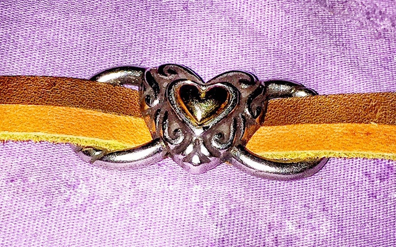 May include: A close-up of a leather bracelet with a heart-shaped clasp. The bracelet features two leather straps in shades of brown and yellow, connected by a silver-colored heart clasp with ornate detailing. The background is a textured purple fabric.