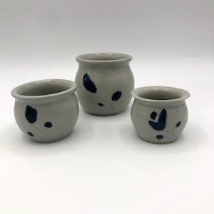 May include: Three small, gray ceramic pots with a rounded shape. Each pot features a dark blue floral design. The pots vary in size, with the largest in the center and the smallest on the right.
