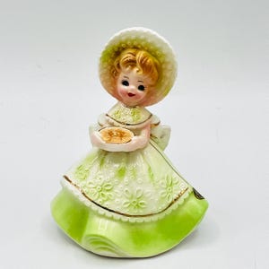 May include: A vintage ceramic figurine of a girl wearing a light green dress and a matching hat. The figurine is holding a small, round object. The dress has a floral pattern and gold trim. The girl has blonde hair and rosy cheeks.