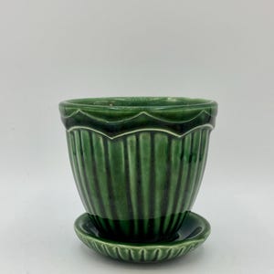 May include: A deep forest green ceramic plant pot with a glossy finish and a matching saucer. The pot features vertical ribbed detailing along the body and a scalloped edge around the top rim, creating a textured appearance for this container.