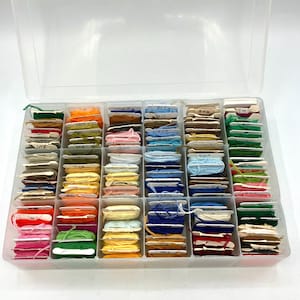 May include: A clear plastic storage box containing numerous spools of embroidery floss in a wide variety of colors. Each spool is wrapped with thread and organized into individual compartments within the rectangular container.