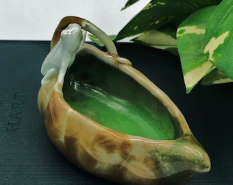 May include: A ceramic boat-shaped dish with a glossy brown and green marbled exterior and a solid bright green interior. A small, light-colored ceramic frog figure is perched on the curved handle, looking into the basin of the vessel.