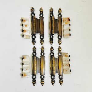 May include: Four antique-style brass hinges with a dark, brushed finish and decorative finials at the top and bottom. Each hinge is accompanied by a small, off-white plastic strip containing four black screws for installation purposes.