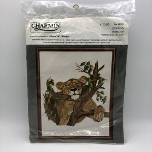 May include: A Charmin Lioness crewel kit. The finished size is 16" x 20". The image shows a detailed embroidery of a lioness resting on a tree branch, with green leaves and brown branches. The kit includes fabric, yarn, floss, needle, and instructions.