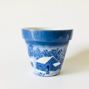 May include: A blue and white ceramic flower pot with a winter scene design. The pot features a blue rim and a detailed illustration of a snow-covered cabin, trees, and the text "IN THE WILDERNESS".
