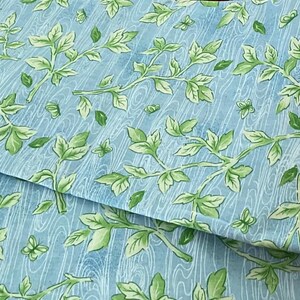 May include: A light blue fabric featuring a printed wood grain texture overlaid with a repeating pattern of green leafy vines. The material is folded, showing the continuous botanical design across the surface of the textile.