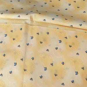 May include: Yellow fabric with a repeating pattern of small blue and yellow butterflies. The fabric has a soft, slightly wrinkled appearance, with subtle white dotted lines. The overall color scheme is a light, warm yellow.