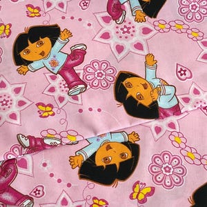 May include: Pink fabric featuring a repeating pattern of a cartoon character in a light blue shirt and pink pants. The design includes pink flowers, butterflies, and star-shaped elements. The fabric is likely intended for children's crafts or apparel.