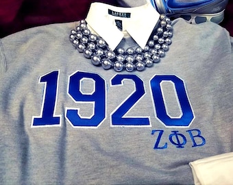 Puede incluir: Una sudadera gris con el número 1920 bordado en azul con un contorno blanco. Las letras ZФB están bordadas en azul debajo del número.