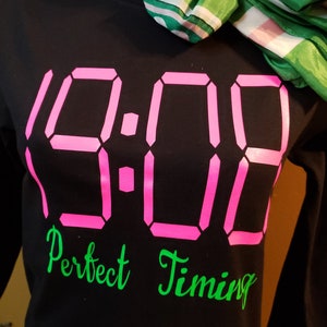 May include: Black long-sleeved shirt with a pink digital clock design that reads "9:00" and the text "Perfect Timing" in green.