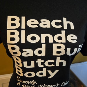 May include: Black t-shirt with white text that reads "Bleach Blonde Bad Built Butch Body" and "Sincerely, A Black Woman's Clap".