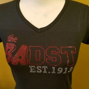 Puede incluir: Camiseta negra con cuello en V con letras de pedrería rojas y blancas. El texto dice "SADST EST.1913". La camiseta tiene un diseño clásico y sencillo.