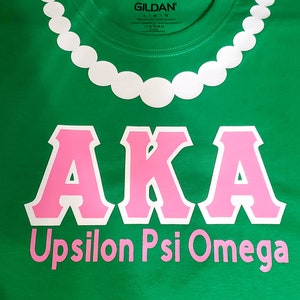 以下が含まれることがあります： ピンク色の「AKA」の文字が白で縁取られ、「Upsilon Psi Omega」の文字がピンク色で描かれた緑色のTシャツ。上部には白いビーズネックレスのデザインがあります。Gildanのロゴが見えます。