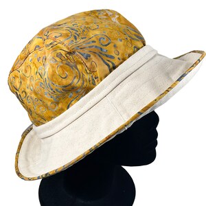 May include: A wide-brimmed sun hat with a patterned crown and a contrasting brim. The crown features a golden yellow batik design with blue and gray swirls. The brim is a light cream color.