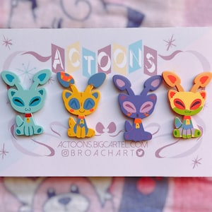 May include: Four colourful cartoon animal pins with a retro design. The pins feature a blue, yellow, purple, and orange alien-like creature with big eyes and a friendly expression. The pins are displayed on a white card with the text "ACTOONS" in a retro font.