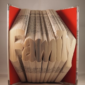 Könnte beinhalten: Ein Buch, das in die Form des Wortes "Familie" gefaltet ist. Das Buch ist geöffnet und die Seiten sind gefaltet, um die Buchstaben zu bilden. Das Buch ist außen rot und die Seiten sind weiß.