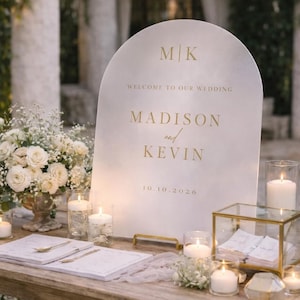Op de afbeelding: Een boogvormig wit welkomstbord voor een bruiloft met gouden letters die M|K, WELCOME TO OUR WEDDING, MADISON and KEVIN en 10.10.2026 weergeven. Het bord staat op een houten tafel naast witte bloemstukken en glazen kaarsen.