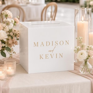 Peut inclure: Une boîte à cartes carrée en acrylique blanc avec une fente horizontale sur le dessus et le texte doré imprimé "MADISON and KEVIN" sur la face avant. La boîte est posée sur une table avec des compositions florales et des bougies allumées.